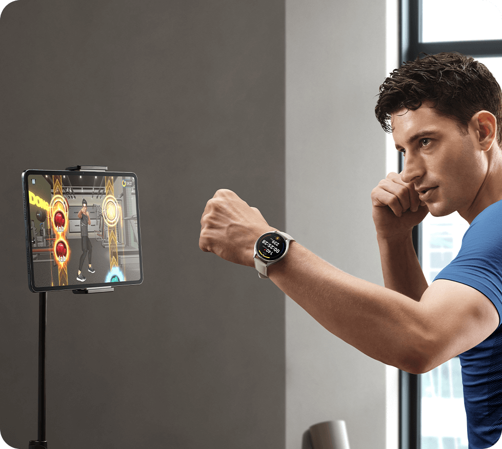 Un homme en t-shirt bleu s'entraîne à la boxe virtuelle en portant une montre connectée Xiaomi Watch 2 à cadran rond et bracelet clair. Il fait face à une tablette fixée sur un support affichant un jeu d'entraînement interactif de fitness.