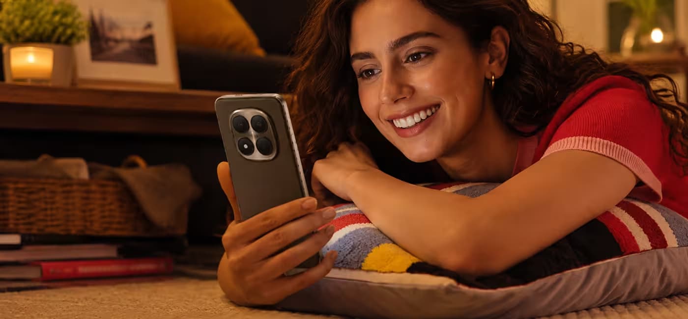 Femme souriante allongée sur un coussin, tenant un smartphone Redmi Note 15 Pro+ 5G dans un intérieur chaleureux et tamisé.